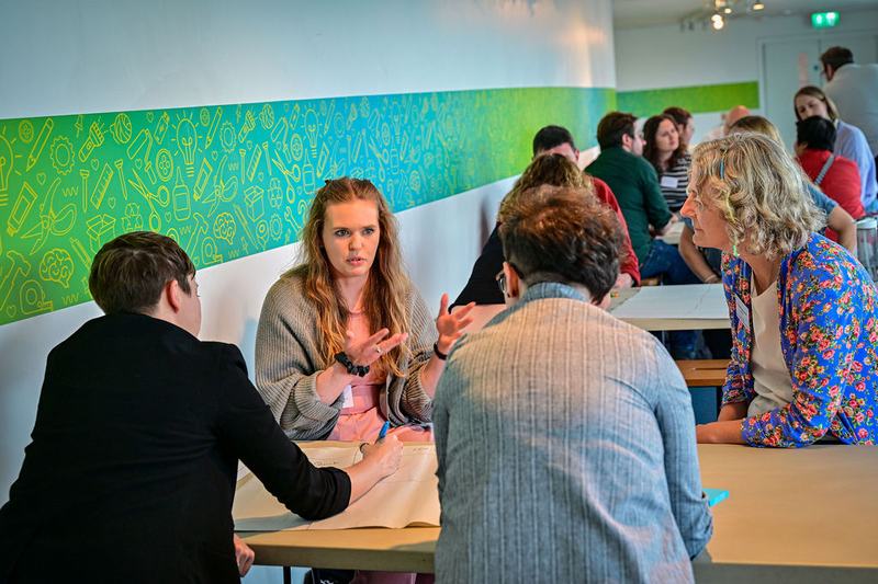 A group of people sit at a table having a discussion, as one writes on a piece of flipchat paper