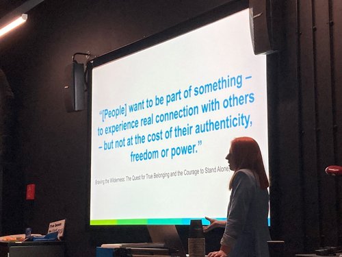 Woman speaking beside a large screen with text "[People want to be part of something - to experience real connection with others - but not at the cost of their authenticity, freedom or power."