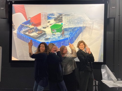 Four women in front of a screen with their hands clasped pointing up to the photo of a messy children&#x27;s activity table